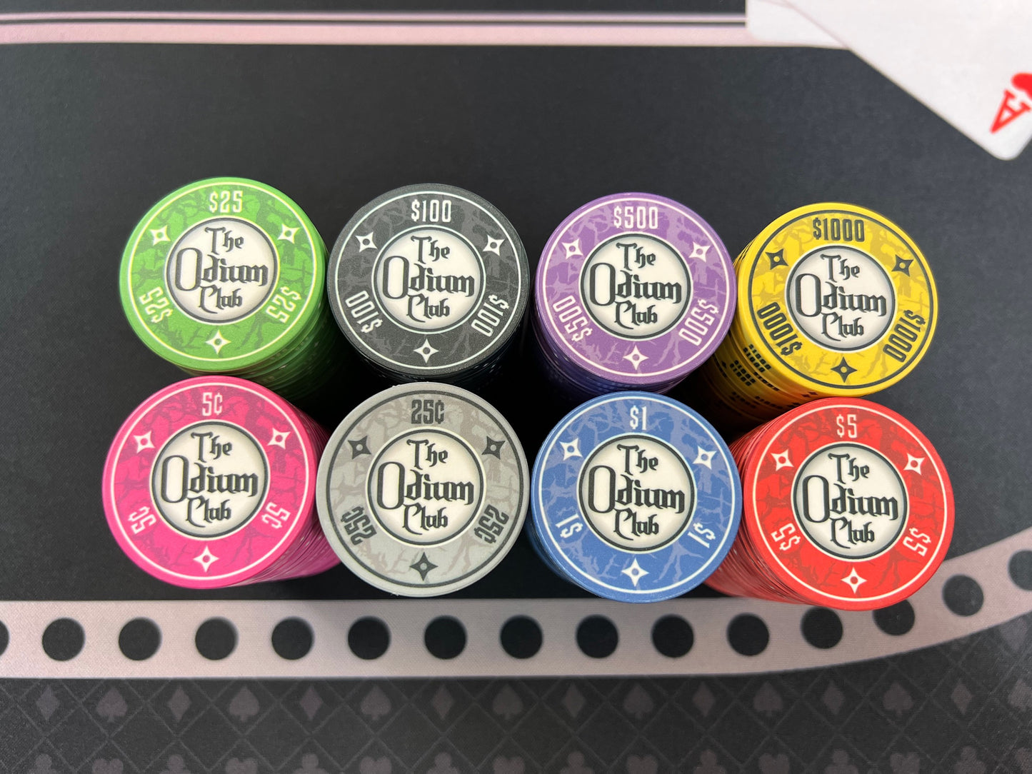 All eight of the stock colors and denominations of the modern-looking Odium Club poker chips, top-down view. Both faces are identical. Each side shows the logo in the poker chip inlay and the poker chip denoms around the edge. Available dollar values are 5¢ pink, 25¢ gray, $1 blue, $5 red, $25 green, $100 black, $500 purple, and $1000 yellow.