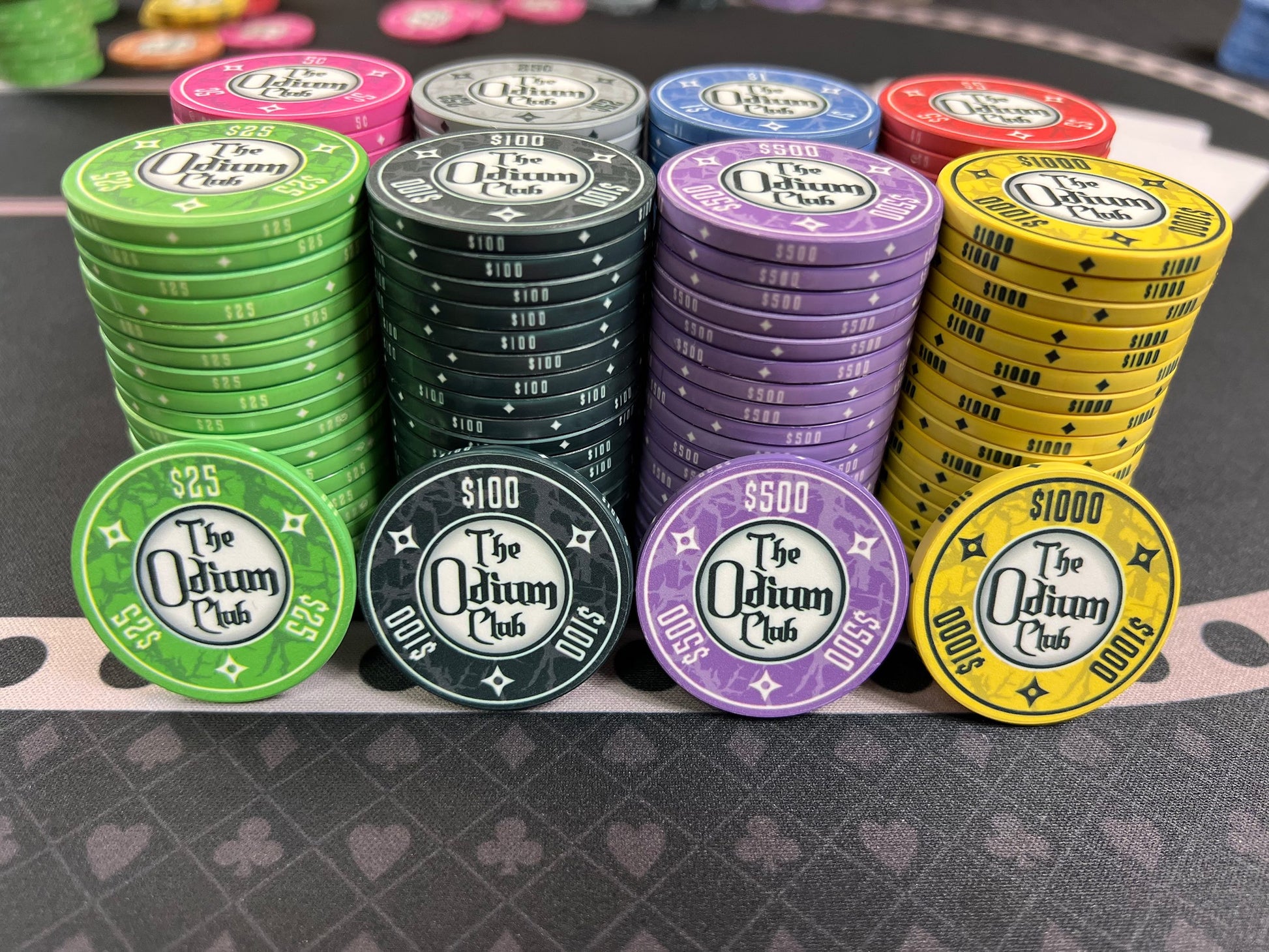 All eight of the stock colors and denominations of the modern-looking Odium Club poker chips in stacks of 20. Both faces are identical. Each side shows the logo and denom. Available in 5¢ pink, 25¢ gray, $1 blue, $5 red, $25 green, $100 black, $500 purple, and $1000 yellow. Shown up front: twenty-five dollar green, one-hundred dollar black, five-hundred dollar purple, and one thousand dollar yellow.