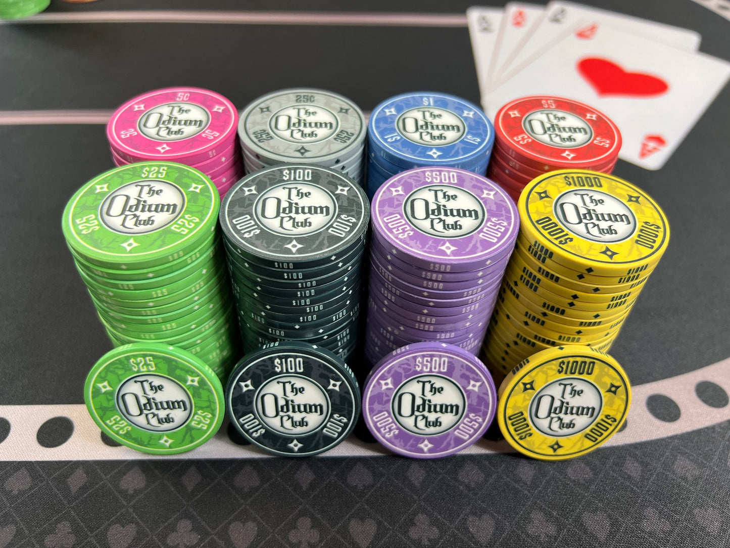 All eight of the stock colors and denominations of the modern-looking Odium Club poker chips in stacks of 20. Both faces are identical. Each side shows the logo and denom. Available in 5¢ pink, 25¢ gray, $1 blue, $5 red, $25 green, $100 black, $500 purple, and $1000 yellow. Shown up front: twenty-five dollar green, one-hundred dollar black, five-hundred dollar purple, and one thousand dollar yellow.